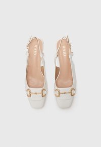 Pari valkoisia slingback-matalakenkiä, joissa on kultainen hevosenpäätyksikko ja neliön muotoinen kärki, esillä valkoista taustaa vasten.