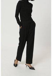 Schwarzes Rollkragenoberteil kombiniert mit hoch taillierten, lässig geschnittenen schwarzen Hosen. Das Outfit zeichnet sich durch eine glatte Textur und ein einfaches Design aus. Schwarze spitze Absätze.