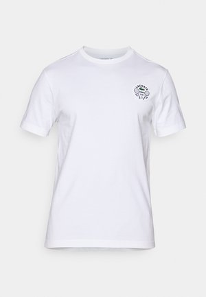 T-shirt en coton blanc avec un col rond et des manches courtes, arborant un logo de club de tennis brodé en noir sur le côté gauche de la poitrine.