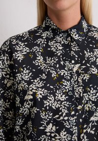 Femme portant une chemise noire à boutons ornée d'un motif floral blanc et vert olive, avec un col et une poche poitrine.