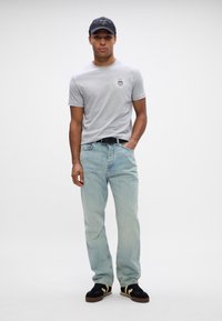Grå bomulds t-shirt med rund halsudskæring og logo, lyseblå jeans, sorte sneakers med beige detaljer og en sort kasket.