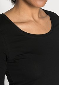 Femme portant un t-shirt noir à encolure dégagée, montrant le cou, la clavicule et le haut de la poitrine sur un fond uni.