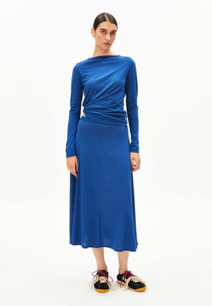 Blauwe jurk met lange mouwen en een gedrapeerd ontwerp, voorzien van een getailleerd lijfje en een vloeiende rok. Gecombineerd met zwarte sneakers met roze veters.