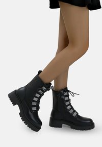 Botas de combate de cuero negro con una suela gruesa, cuentan con una cremallera lateral y detalles en panel de cordones plateados para añadir textura y estilo.
