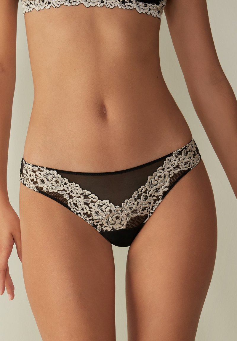 Ensemble de lingerie en dentelle noire présentant une délicate broderie florale le long de la taille et un panneau transparent, offrant un design moderne et élégant.