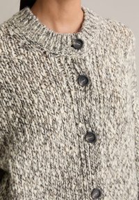 Cárdigan de punto grueso en tonos crema y gris, con un cuello alto, cinco botones negros y una tela texturizada y tejida.