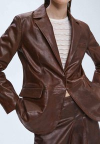 Ruskea nahkatakki, jossa on sileä pinta, istuva muoto, kaksi etutaskua ja yksi napin sulkija. Puettuna vaalean neulepaidan päällä.