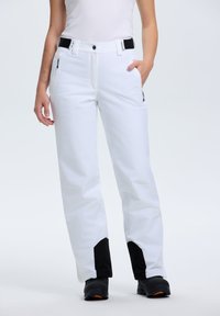 Pantaloni da sci bianchi con una texture liscia, vita regolabile, tasche laterali con zip e dettagli neri sui polsini. Indossati con stivali neri.