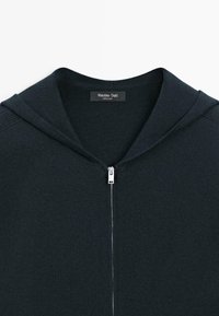 Ciemnoszara bluza z kapturem zapinana na zamek, wykonana z teksturowanej tkaniny. Posiada kaptur i srebrny zamek błyskawiczny. Wewnątrz przy szyi widoczna metka.