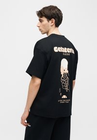 Νεαρός άνδρας με μαύρο μπλουζάκι που έχει εκτυπωμένο ένα μπεζ χωνάκι παγωτού και το κείμενο «Gelateria Ellesse» στην πλάτη, κοιτάζοντας πλάγια σε λευκό φόντο.