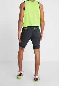 Man som bär en neongrön ärmlös mesh-top, svarta träningsshorts med gröna och orange detaljer, och vita löparskor med neongröna sulor.