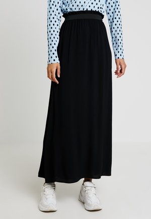 Kvinde iført en sort, høj taljet lang nederdel kombineret med hvide sneakers og en blå langærmet bluse med sorte polkaprikker.