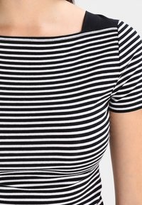 Haut rayé noir et blanc avec un décolleté carré, des manches courtes et une texture en coton lisse. Comprend des accents noirs solides au niveau des épaules.