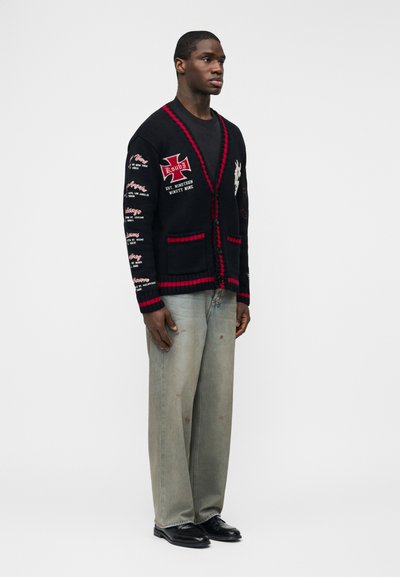 Άνδρας στέκεται φορώντας μαύρη ζακέτα με κόκκινες λεπτομέρειες και λευκά κεντήματα με κείμενο, ξεθωριασμένο τζιν με φαρδιές παντελόνες, μαύρα παπούτσια, σε απλό λευκό φόντο.