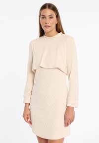 Beiges Kleid mit einem strukturierten, gesteppte Rock und einem glatten, mehrlagigen Oberteil. Hat einen hohen Ausschnitt und lange Ärmel.