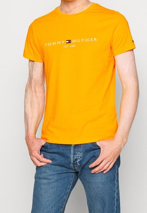 Pomarańczowa koszulka z krótkim rękawem wykonana z bawełny, z białym logo na środku i granatowym akcentem na rękawie. Klasyczny krój z okrągłym dekoltem.