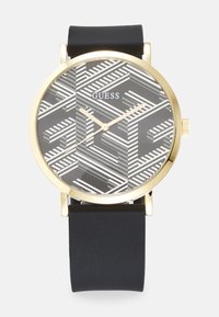 Goldfarbene Uhr mit einem schwarzen Silikonarmband, ausgestattet mit einem grau gemusterten Zifferblatt mit weißen Streifen und dem Markennamen "GUESS".