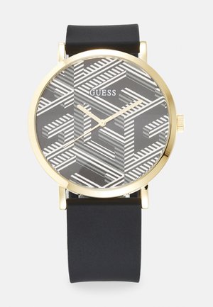 Orologio dorato con cinturino in silicone nero, dotato di un quadrante grigio a motivo geometrico con strisce bianche e il nome del marchio "GUESS" in evidenza.