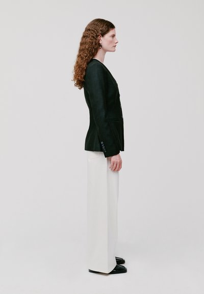 Blazer in lino nero con dettagli in bottone, abbinato a pantaloni bianchi a gamba larga e scarpe nere. Il modello è in profilo contro uno sfondo neutro.