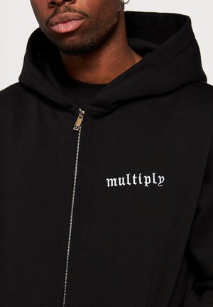 Homme portant un sweat à capuche zippé noir avec la capuche relevée, avec un texte blanc "multiply" sur la poitrine.