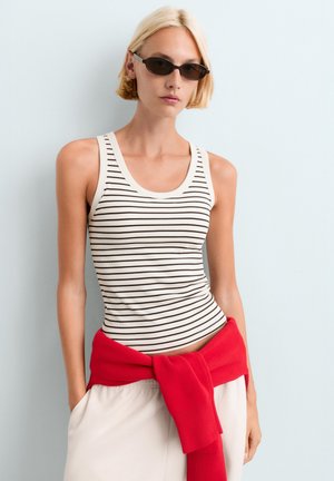 Femme blonde portant des lunettes de soleil noires, un débardeur blanc à rayures noires, un pantalon blanc et un pull rouge noué autour de sa taille.