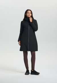 Manteau imperméable noir avec fermeture éclair à l'avant, col haut et deux poches latérales, associé à des leggings marron et des chaussures de sport noires.