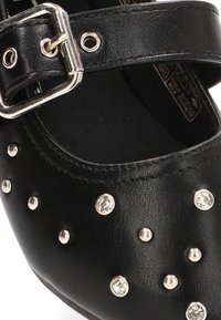 Zapato de cuero negro con correa y hebilla de plata, adornado con remaches metálicos y piedras de strass en la zona de los dedos. Textura suave.