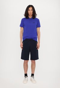 Polo shirt blu royal con logo nero, abbinato a shorts blu navy fino al ginocchio. Il modello indossa sneakers bianche e calze nere alla caviglia.