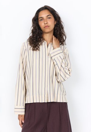 Woman with curly hair wearing a loose beige shirt with vertical stripes and dark high-waisted pants, standing with one hand on her neck.