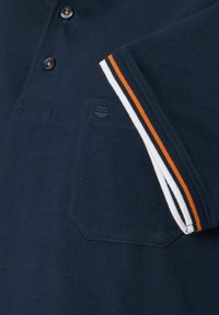 Granatowa koszulka polo z trzema guzikami, kieszonką na piersi oraz paskowanymi mankietami w białym i pomarańczowym kolorze. Gładka tekstura bawełny.