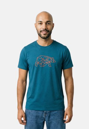 Man met baard die een blauw t-shirt draagt met een geometrisch berenontwerp en blauwe spijkerbroek, staand tegen een egale lichte achtergrond.