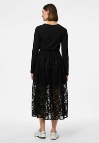 Haut à manches longues noir associé à une jupe maxi en dentelle noire transparente. Présente des motifs floraux en dentelle et une taille élastique. Porté avec des baskets argentées.