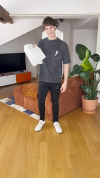 Hallhalli hallitatud t-särk logo ja mustad kulunud teksad ning valged spordijalatsid. Hoides volditud heledat eset toas, kus on diivan ja taimed.