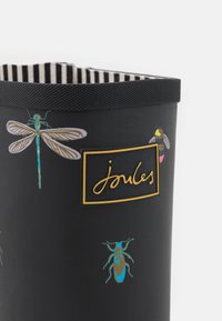 Botte en caoutchouc noire, imperméable, ornée d'illustrations d'insectes colorés, avec un logo doré et une doublure intérieure rayée. Bord supérieur texturé noir.