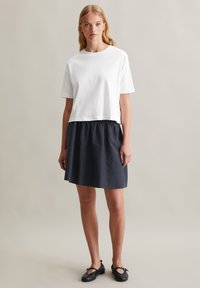 T-shirt blanc raccourci associé à une jupe bleue marine froncée. La jupe a un tissu texturé et tombe au-dessus des genoux. Des chaussures plates noires complètent la tenue.