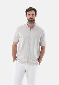 Camicetta grigia chiara a maniche corte con bottoni, realizzata in tessuto morbido, con colletto e chiusura frontale a bottoni. Abbinala a pantaloni bianchi.