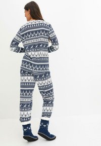 Une combinaison à motifs bleu marine et blanc, présentant des flocons de neige, des rennes et des arbres ; en tissu doux ; coupe décontractée ; assortie à des bottes en polaire bleu marine.