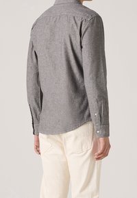 Camicia grigia a maniche lunghe con colletto button-down, tessuto morbido, orlo curvo e polsini bottonati, abbinata a pantaloni chiari.