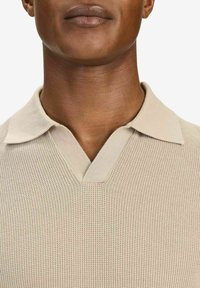 Beige stickad polo-shirt med ett texturerat mönster, med V-hals och krage. Närbild av överkroppen.