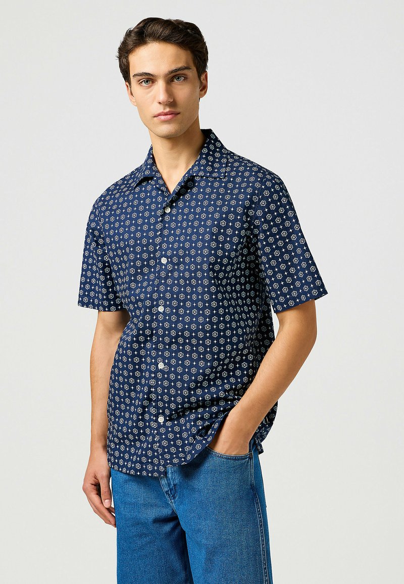 Camisa de manga corta azul marino con un patrón floral y acentos en blanco. Presenta un collar y un cierre frontal de botones, fabricada con un tejido ligero.