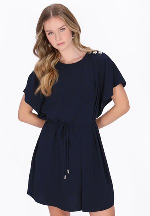 Femme aux longs cheveux ondulés portant une robe bleu marine ceinturée avec des détails de boutons sur l'épaule, posant les mains derrière le dos sur un fond blanc.