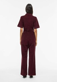 Tuta in velluto a coste bordeaux con maniche corte, vita con cintura, design a gamba larga e tasche posteriori. Texture liscia e vestibilità sartoriale.