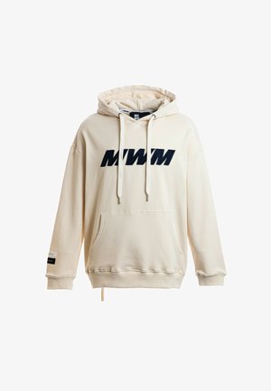 Persona joven con cabello rubio lleva una sudadera con capucha color crema, pantalones cortos deportivos blancos con rayas de colores, calcetines blancos y zapatos beige texturizados sin cordones.