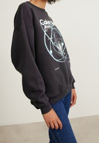 Černá mikina s kulatým výstřihem, volným oversized střihem, s grafickým potiskem v modré a bílé barvě, s žebrovanými manžetami a lemem. Nošena s modrými džínami.