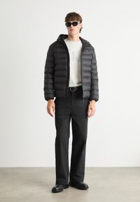 Giacca a vento nera con cappuccio, cuciture orizzontali, zip frontale, indossata sopra una maglietta grigio chiaro. I pantaloni e le scarpe nere completano il look.
