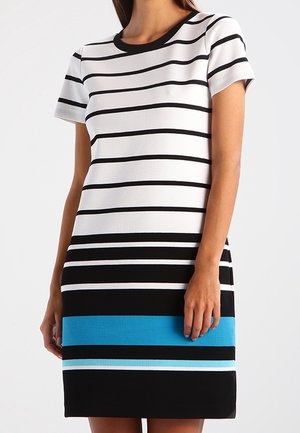 Kvinde iført en kortærmet, knælængde kjole med horisontale sorte, hvide og blå striber på en ensfarvet baggrund.