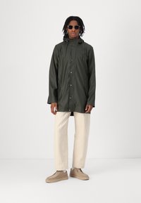 Groene waterdichte jas met capuchon, voorpand met drukknoopsluiting en twee zijzakken, gecombineerd met crèmekleurige broeken en tan kleurige schoenen.