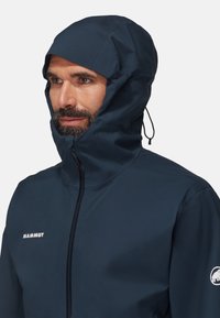 Veste Mammut bleu marine avec un col haut et une capuche ajustable, dotée d'une fermeture éclair sur le devant et d'un logo sur la partie gauche de la poitrine.