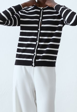 Personne portant un cardigan rayé horizontal noir et blanc avec des boutons en perles et un pantalon blanc, ajustant le col.