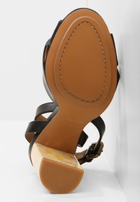 Sandale noire à brides avec un talon carré bloc présentant un motif à carreaux et une semelle cousue de couleur marron, positionnée à la verticale contre une surface blanche.
